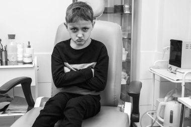 Siyah ve beyaz fotoğraf, küçük bir çocuk, sarışın bir genç, otolaringolojistin ofisinde oturmuş, incelenmeyi bekliyor, endişeli, duygusal, ama gülümseyen, sağlık hizmetleri. Yazmak için bir yer