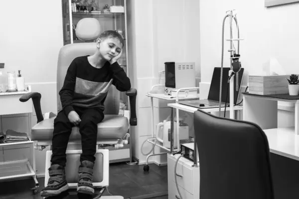Black and white photo, a small, fair-haired teenager, sitting in the otolaryngologist\'s office for an examination, afraid, thoughtful. Health care
