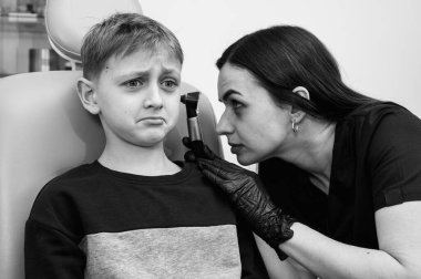 Siyah ve beyaz fotoğraf tıbbi muayenesi, küçük sarışın çocuk, genç, otolaryngolog ofis koltuğunda oturan doktor kulağını inceliyor, endişeli, duygusal, ama gülümseyen.