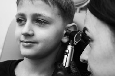 Siyah ve beyaz fotoğraf tıbbi muayenesi, küçük sarışın çocuk, genç, otolaryngolog ofis koltuğunda oturan doktor kulağını inceliyor, endişeli, duygusal, ama gülümseyen.