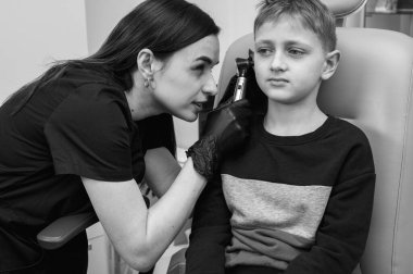 Siyah ve beyaz fotoğraf tıbbi muayenesi, küçük sarışın çocuk, genç, otolaryngolog ofis koltuğunda oturan doktor kulağını inceliyor, endişeli, duygusal, ama gülümseyen.