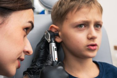 Klinikte otoskoplu bir doktor tarafından muayene edilen, sandalyede oturan, endişeli, sarışın bir çocuğun portresi. Görüntüyü kapat