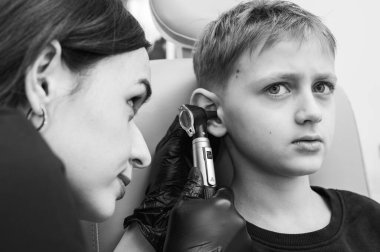 Siyah ve beyaz, sarışın bir çocuğun portresi klinikte otoskoplu bir doktor tarafından inceleniyor, bir sandalyede oturuyor, endişeli. Görüntüyü kapat