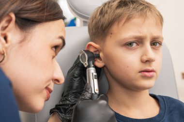 Klinikte otoskoplu bir doktor tarafından muayene edilen, sandalyede oturan, endişeli, sarışın bir çocuğun portresi. Görüntüyü kapat