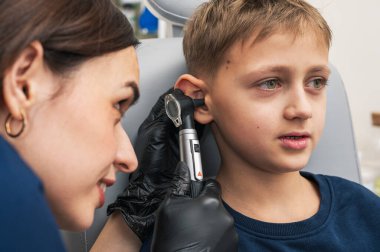 Küçük bir çocuk, sarışın, gülümseyen, otolaryngolojistin ofisinde oturan, doktorlara güvenen, duygusal, gülümseyen kulakları inceleyen. Fiziksel muayene.