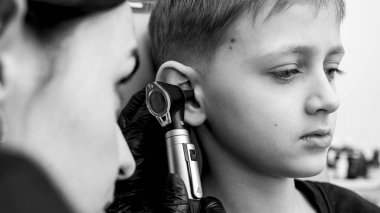 Siyah ve beyaz, sarışın bir çocuğun portresi klinikte otoskoplu bir doktor tarafından inceleniyor, bir sandalyede oturuyor, endişeli. Görüntüyü kapat