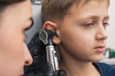 Küçük bir çocuk, sarışın, gülümseyen, otolaryngolojistin ofisinde oturan, doktorlara güvenen, duygusal, gülümseyen kulakları inceleyen. Fiziksel muayene.