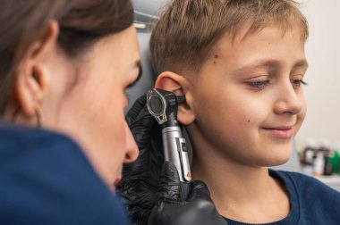 Küçük bir çocuk, sarışın, gülümseyen, otolaryngolojistin ofisinde oturan, doktorlara güvenen, duygusal, gülümseyen kulakları inceleyen. Fiziksel muayene.