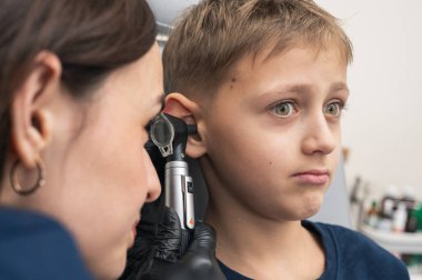 Klinikte otoskoplu bir doktor tarafından muayene edilen, sandalyede oturan, endişeli, sarışın bir çocuğun portresi. Görüntüyü kapat