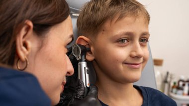 Küçük bir çocuk, sarışın, gülümseyen, otolaryngolojistin ofisinde oturan, doktorlara güvenen, duygusal, gülümseyen kulakları inceleyen. Fiziksel muayene.