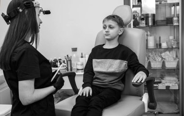 siyah ve beyaz fotoğraf, boğaz muayenesi, küçük bir çocuk, sarışın bir genç, otolaringolojistin ofisinde oturuyor, doktor boğazını inceliyor, endişeli, duygusal, ama gülümsüyor. Fiziksel muayene.