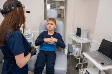  Küçük bir çocuk, sarışın bir genç, otolaringolojistin ofisinde oturuyor, doktorda kulak muayenesi yaptırıyor, endişeli, duygusal, ama gülümsüyor. Fiziksel muayene.