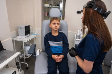  Küçük bir çocuk, sarışın bir genç, otolaringolojistin ofisinde oturuyor, doktorda kulak muayenesi yaptırıyor, endişeli, duygusal, ama gülümsüyor. Fiziksel muayene.