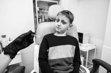 siyah ve beyaz fotoğraf, boğaz muayenesi, küçük bir çocuk, sarışın bir genç, otolaringolojistin ofisinde oturuyor, doktor boğazını inceliyor, endişeli, duygusal, ama gülümsüyor. Fiziksel muayene.