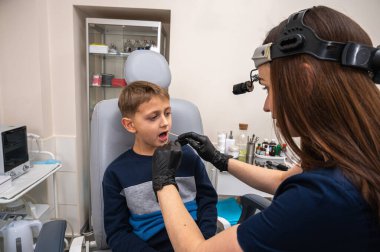 Küçük bir çocuk, sarışın bir genç, otolaryngologun ofisinde oturuyor, muayene ediliyor, boğazından örnek alıyor, doktor tarafından test ediliyor, duygusal ama gülümsüyor. Fiziksel muayene.
