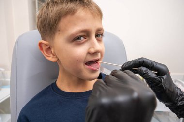 Küçük bir çocuk, sarışın bir genç, otolaryngologun ofisinde oturuyor, muayene ediliyor, boğazından örnek alıyor, doktor tarafından test ediliyor, duygusal ama gülümsüyor. Fiziksel muayene.