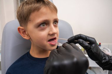 Boğaz muayenesi, küçük bir çocuk, sarışın bir genç, otolaringolojistin ofisinde oturuyor, doktor boğazını inceliyor, endişeli, duygusal, ama gülümsüyor. Fiziksel muayene.