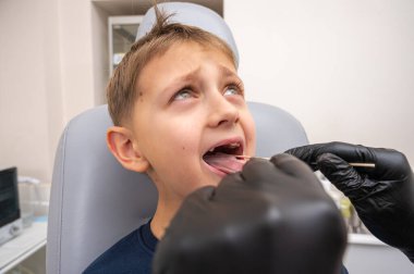 Küçük bir çocuk, sarışın bir genç, otolaryngologun ofisinde oturuyor, muayene ediliyor, boğazından örnek alıyor, doktor tarafından test ediliyor, duygusal ama gülümsüyor. Fiziksel muayene.