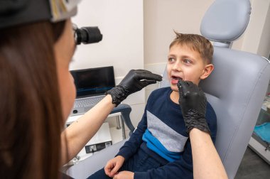 Küçük bir çocuk, sarışın bir genç, otolaryngologun ofisinde oturuyor, muayene ediliyor, boğazından örnek alıyor, doktor tarafından test ediliyor, duygusal ama gülümsüyor. Fiziksel muayene.