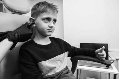 Siyah beyaz bir fotoğraf, küçük bir çocuk, sarışın bir genç, bir otolargologun ofisinde oturuyor, doktorun kulaklarını inceliyor, endişeli, duygusal, ama gülümsüyor. Fiziksel muayene.