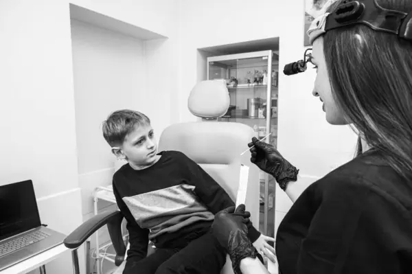 black and white photo, throat examination, little boy, fair-haired teenager, sitting in otolaryngologist\'s office, doctor examines throat, worried, emotional, but smiling. Physical examination
