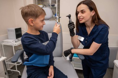 Küçük bir çocuk, sarışın bir genç, otolaryngolojistin ofisinde, doktorun kulak muayenesinde, doktorla oynuyor, duygusal ama gülümseyen, rol yapan. Fiziksel muayene.