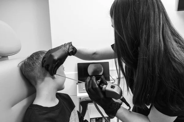 black and white photo, little boy, fair-haired teenager, sitting in an otolaryngologist\'s office, examining the doctor\'s ears, worried, emotional, but smiling. Physical examination
