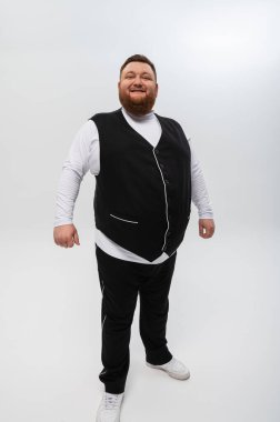 Beyaz arka planda siyah yelek ve açık tişörtlü mutlu sakallı bir adamın duygusal portresi. Stüdyo çekimi. Duygular, yazacak bir yer.