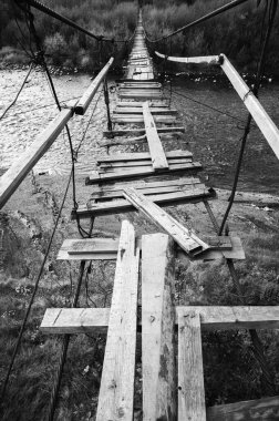 siyah ve beyaz fotoğraf, eski bir asma köprü nehir boyunca uzanıyor, macera ve tehlike hissi yaratıyor. Yıpranmış ahşap tahtalar bir risk atmosferi ve resimli kırsal manzaralar ekler.