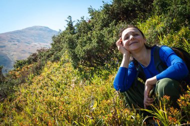 Sırt çantalı dağlarda parlak yapraklarla çevrili yemyeşil ormanda doğa yürüyüşünden zevk alan gülümseyen bir kadın. Açık hava macerası ve doğal çevrede sükunet, dağlar.