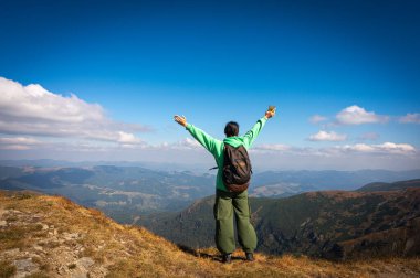 Turist, yeşil pantolonlu bir kız ve sırt çantalı bir kazak zirveyi fethetmeyi, kollarını kaldırmayı, parlak mavi ışık altında dağların panoramik manzarasının keyfini çıkarmayı, arkadan manzarayı kutluyor.