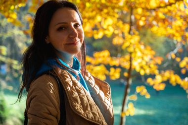 Altın yapraklarla çevrili yumuşak sonbahar güneşinde gülümseyen bir kadının portresi. Sonbahar mevsiminin neşe ve güzelliğini yansıtan sıcak ve canlı bir açık hava sahnesi.