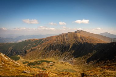 Yeşil bitki örtüsü ve kayalık dokularla kaplı yumuşak güneş ışığıyla yıkanmış dağ yamaçlarının manzarası. Yüksek rakımlı arazinin engebeli güzelliğini gösteren huzurlu bir doğal manzara.