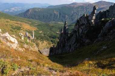 Yeşil bitki örtüsü ve kayalık dokularla kaplı yumuşak güneş ışığıyla yıkanmış dağ yamaçlarının manzarası. Yüksek rakımlı arazinin engebeli güzelliğini gösteren huzurlu bir doğal manzara.