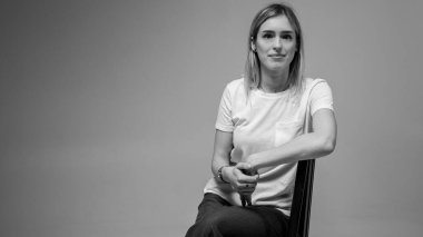 Siyah ve beyaz fotoğraf, kendine güvenen genç bir kız bir sandalyede oturuyor, beyaz bir tişört giyiyor, yana bakıyor. Tarafsız bir geçmişe sahip modern minimalist portre, iş veya hayat konuları için, yansımalar
