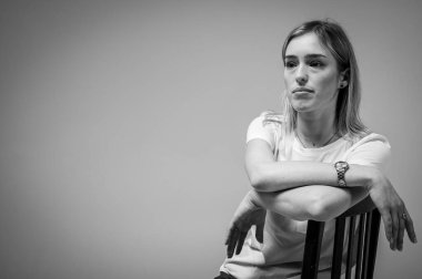 Siyah ve beyaz fotoğraf, kendine güvenen genç bir kız bir sandalyede oturuyor, beyaz bir tişört giyiyor, yana bakıyor. Tarafsız bir geçmişe sahip modern minimalist portre, iş veya hayat konuları için, yansımalar