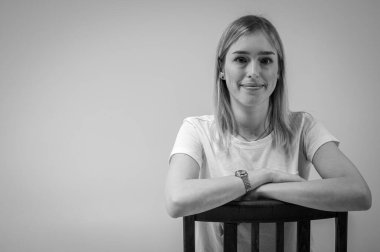 Siyah ve beyaz fotoğraf, kendine güvenen genç bir kız bir sandalyede oturuyor, beyaz bir tişört giyiyor, yana bakıyor. Tarafsız bir geçmişe sahip modern minimalist portre, iş veya hayat konuları için, yansımalar
