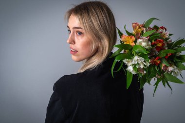 Elegant blonde woman in a black outfit holding a bouquet of colorful flowers over her shoulder. Studio portrait with soft lighting, beauty, fashion, and floral concept clipart