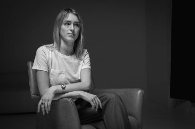 siyah ve beyaz fotoğraf, genç, duygusal, iş kadını deri bir sandalyede oturan, beyaz tişört giyen, stres, endişeler, dosdoğru ileriye bakan. Modern minimalist iş portresi ya da hayat.