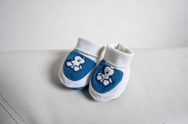 Beyaz ve mavi renkli şirin oyuncak ayı işlemeli şirin bir çift bebek paçası, beyaz deri bir yüzeye yerleştirilmiş. Yumuşak yeni doğmuş ayakkabılar, bebek modası, bebek malzemeleri.