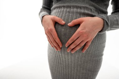 Gri elbiseli hamile bir kadın karnına yakın bir yerde ellerini tutuyor ve bebek bekliyor. Annelik, hamilelik, doğum sonrası bakımı ve bebek beklemenin sembolü. Stüdyo görüntüsü, yakın plan görüntü
