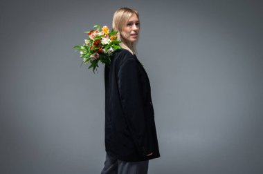 Tarz sahibi, siyah takım elbiseli genç sarışın kız omzunda bir buket taze çiçek tutuyor, arkadan bakıyor, uzun boylu. Modern, zarif ve modaya uygun bir stüdyo portresi.