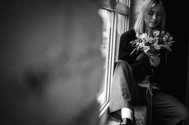 Siyah beyaz fotoğraf, pencerenin kenarında oturan düşünceli genç kız, elinde güzel bir buket çiçek, bahar, renkli. Sıcak atmosfer, doğal ışıklandırma ve romantik ruh hali. Mutluluk ve barış kavramı, beklentiler ve deneyimler