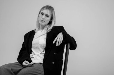 Siyah beyaz fotoğraf, şık bir iş elbisesi içinde gülümseyen genç kız tarafsız bir arka planda bir taburede oturuyor, yorgun, düşünceli. Özgüvenli ve modaya uygun, modern moda portresi, metin için alan