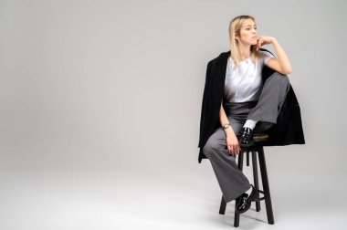 Tarafsız arka planda taburede oturan şık iş kıyafeti içinde gülümseyen genç bir kadın. Kendine güvenen ve modaya uygun, modern moda portresi, tam boy, metin için alan