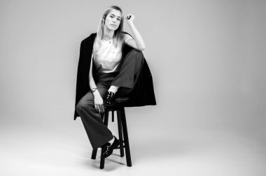 Siyah beyaz fotoğraf, şık bir iş elbisesi içinde gülümseyen genç kız tarafsız bir arka planda bir taburede oturuyor, yorgun, düşünceli. Özgüvenli ve modaya uygun, modern moda portresi, metin için alan