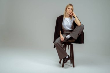 Tarafsız arka planda taburede oturan şık iş kıyafeti içinde gülümseyen genç bir kadın. Kendine güvenen ve modaya uygun, modern moda portresi, tam boy, metin için alan