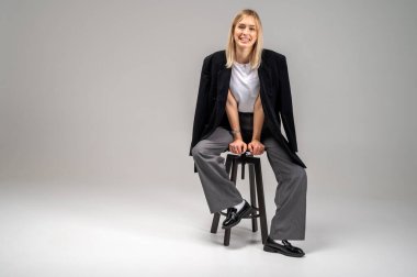 Tarafsız arka planda taburede oturan şık iş kıyafeti içinde gülümseyen genç bir kadın. Kendine güvenen ve modaya uygun, modern moda portresi, tam boy, metin için alan