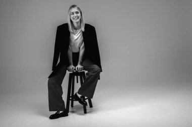 Siyah beyaz fotoğraf, şık bir iş elbisesi içinde gülümseyen genç kız tarafsız bir arka planda bir taburede oturuyor, yorgun, düşünceli. Özgüvenli ve modaya uygun, modern moda portresi, metin için alan