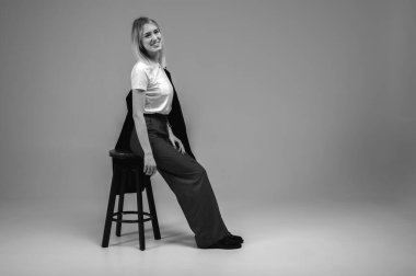 Siyah beyaz fotoğraf, şık bir iş elbisesi içinde gülümseyen genç kız tarafsız bir arka planda bir taburede oturuyor, yorgun, düşünceli. Özgüvenli ve modaya uygun, modern moda portresi, metin için alan